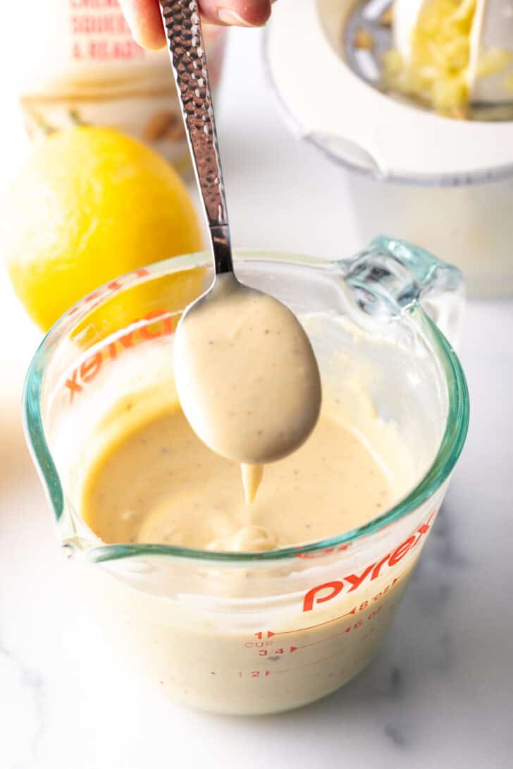 Spoonful of dressing held above a glass measuring cup of tahini dressing.