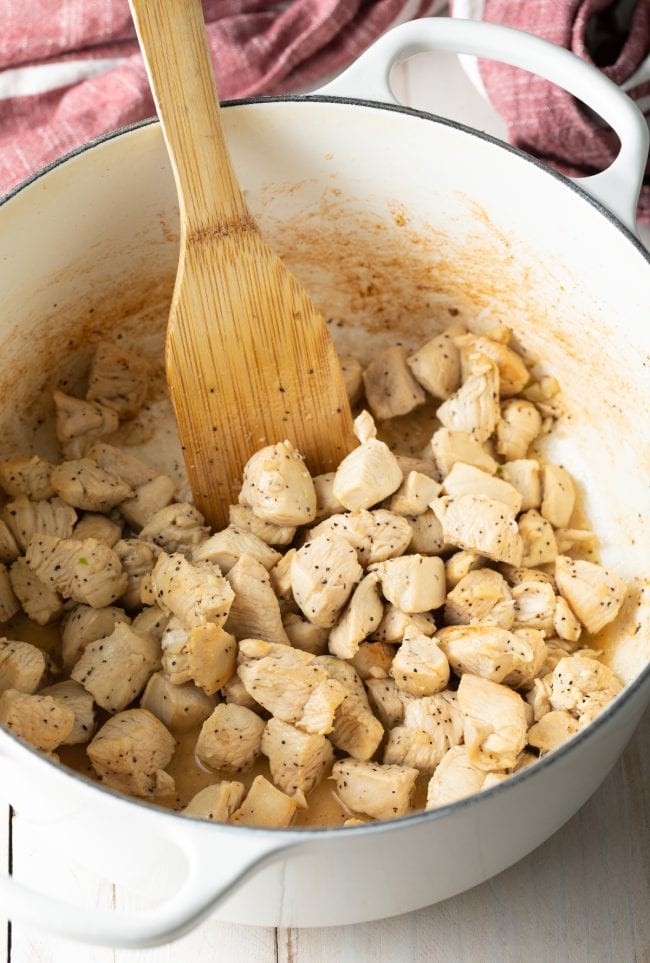 Sautéing chunks of chicken in a white dutch oven with a wood spatula.