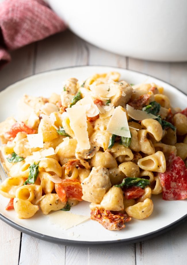 White plate with a serving of creamy chicken spinach pasta with red peppers, sun dried tomatoes, and topped with shaved parmesan cheese.