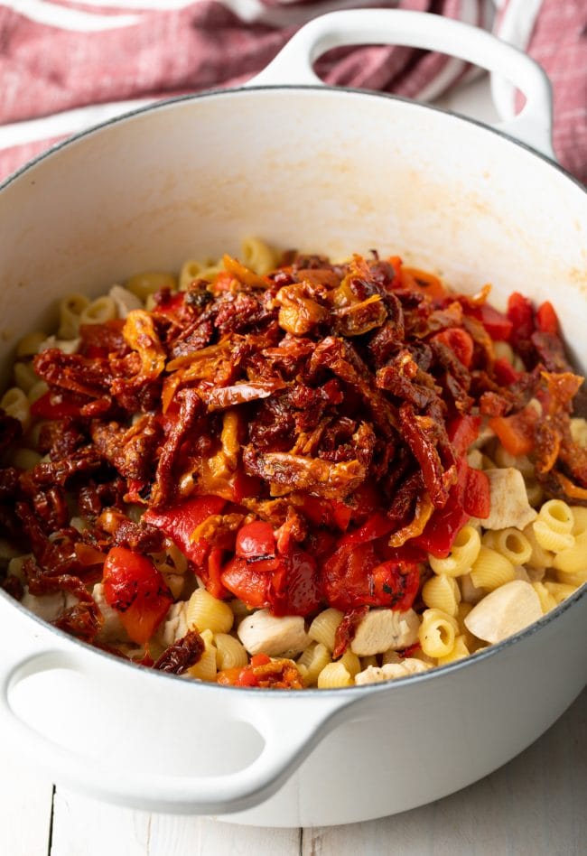 White cooking pot with chunks of cooked chicken and raw macaroni pasta noodles, topped with roasted red peppers and sun dried tomatoes.