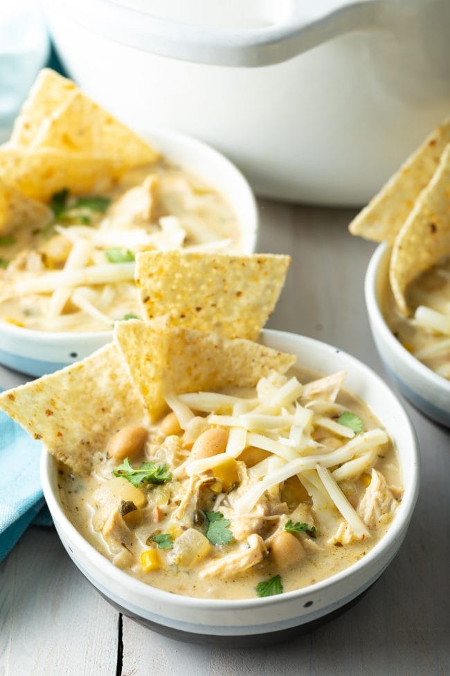 creamy white chicken chili recipe in blue and white bowls with tortilla chips and cheese on top