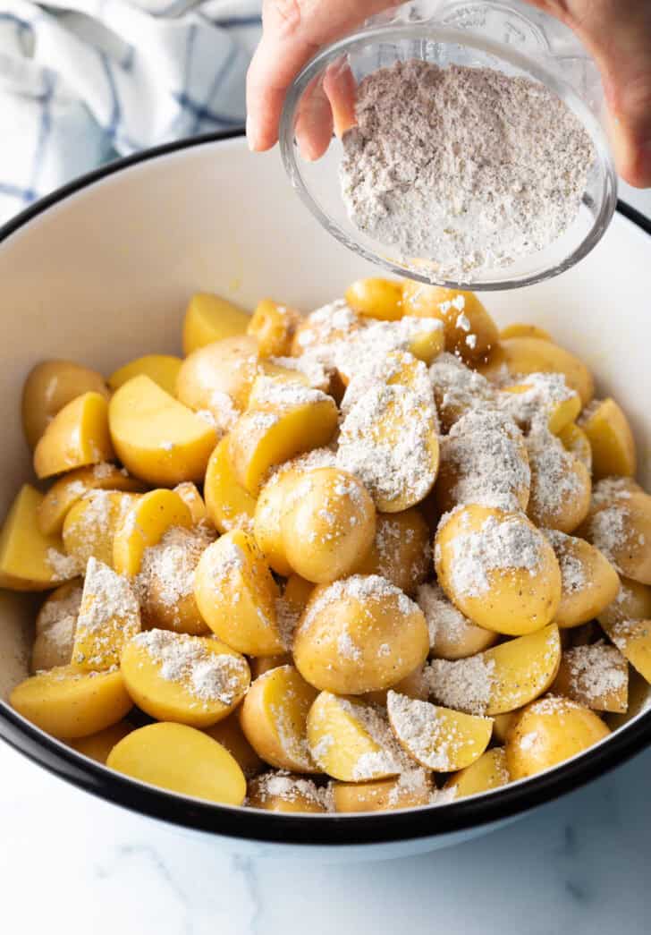 Adding cornstarch and seasoning to a large white bowl of halved yellow potatoes.