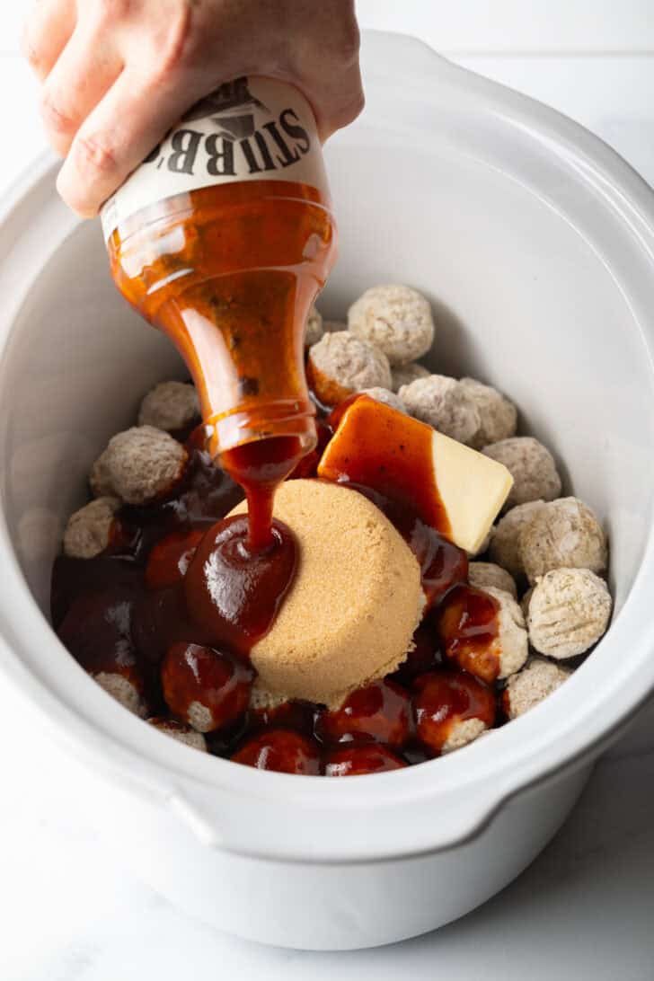 Crockpot filled with frozen meatballs, a stick of butter, scoop of brown sugar, and pouring in a bottle of bbq sauce.