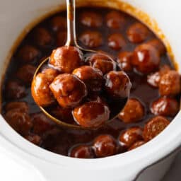 White slow cooker filled with meatballs in bbq sauce. A ladle full of meatballs in being held to camera.