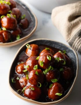 Bowl full of meatballs covered in barbecue sauce, topped with fresh chopped scallions.