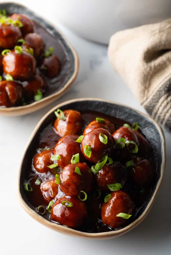 Bowl full of meatballs covered in barbecue sauce, topped with fresh chopped scallions.