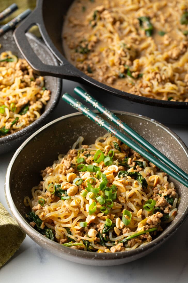 Spicy Szechuan noodles in a black bowl topped with green onion slices. Set of chopsticks are resting on top of the bowl.