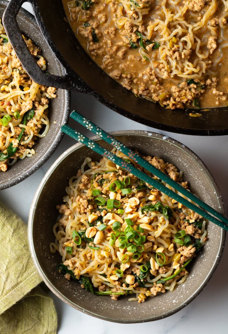 Spicy Szechuan noodles in a black bowl topped with green onion slices. Set of chopsticks are resting on top of the bowl.