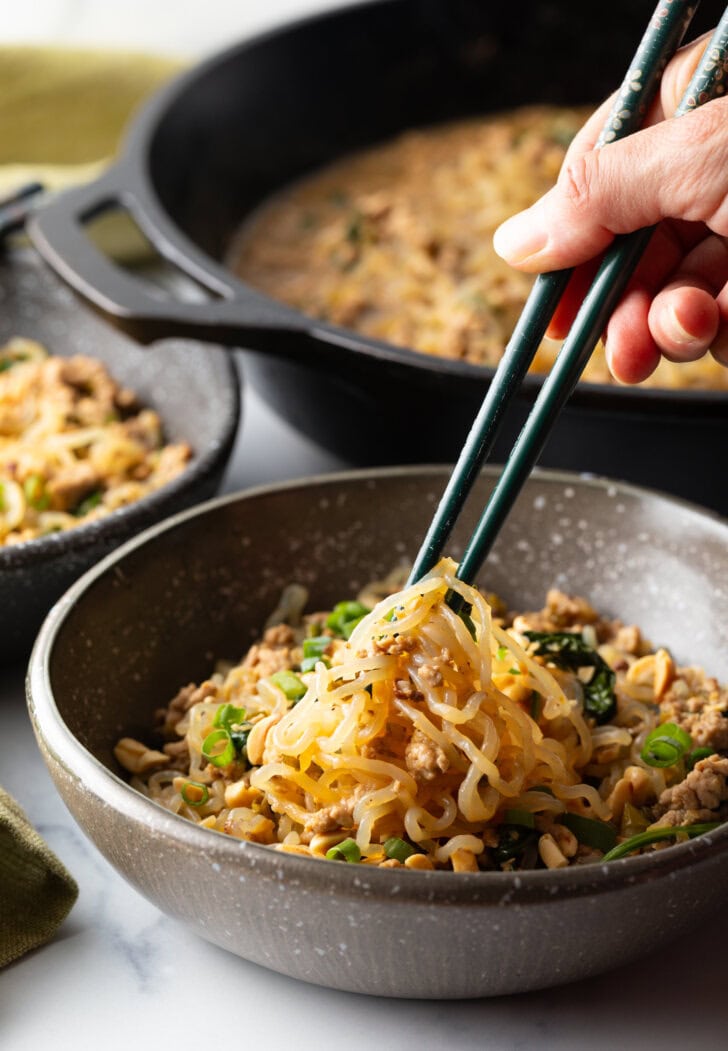 Spicy Szechuan noodles in a black bowl topped with green onion slices. Hand with set of chopsticks are grabbing a bite of noodle.