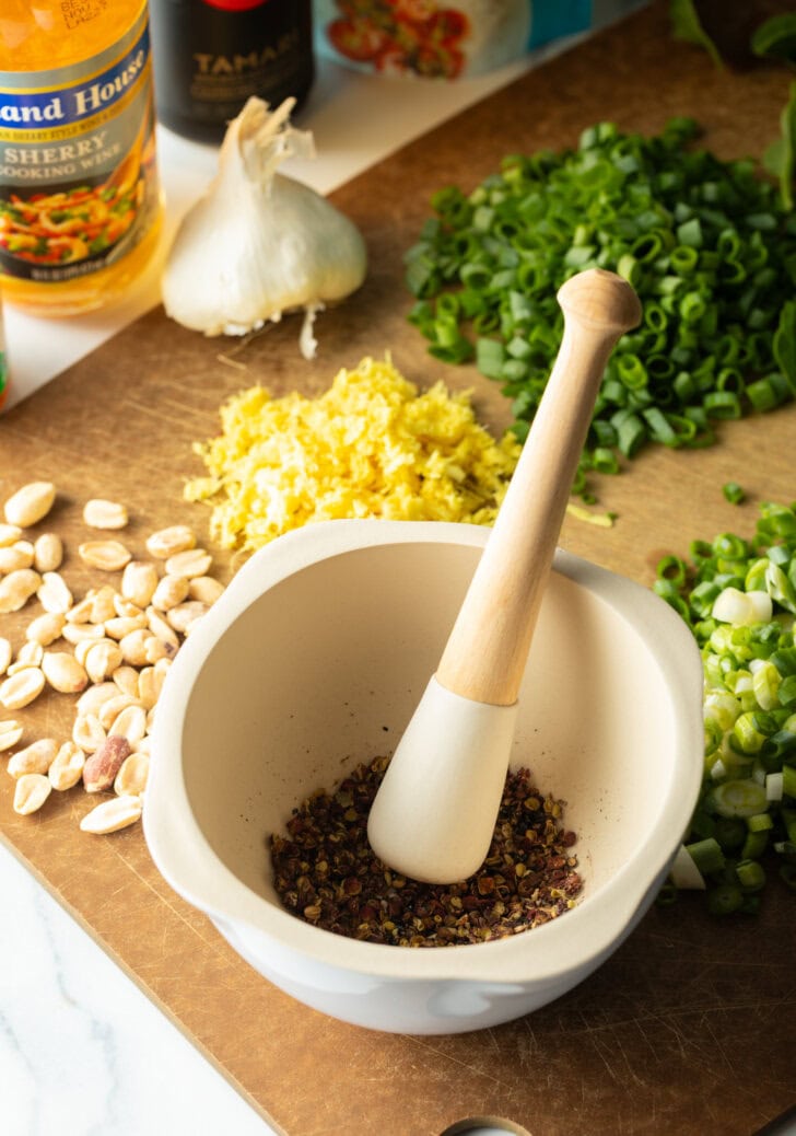 Recipe ingredients: Peppercorns with a mortal and pestel, cutting board with piles of sliced garlic, minced ginger, sliced onions, and spinach.