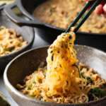 Spicy Szechuan noodles in a black bowl topped with green onion slices. Hand with set of chopsticks are grabbing a bite of noodle.