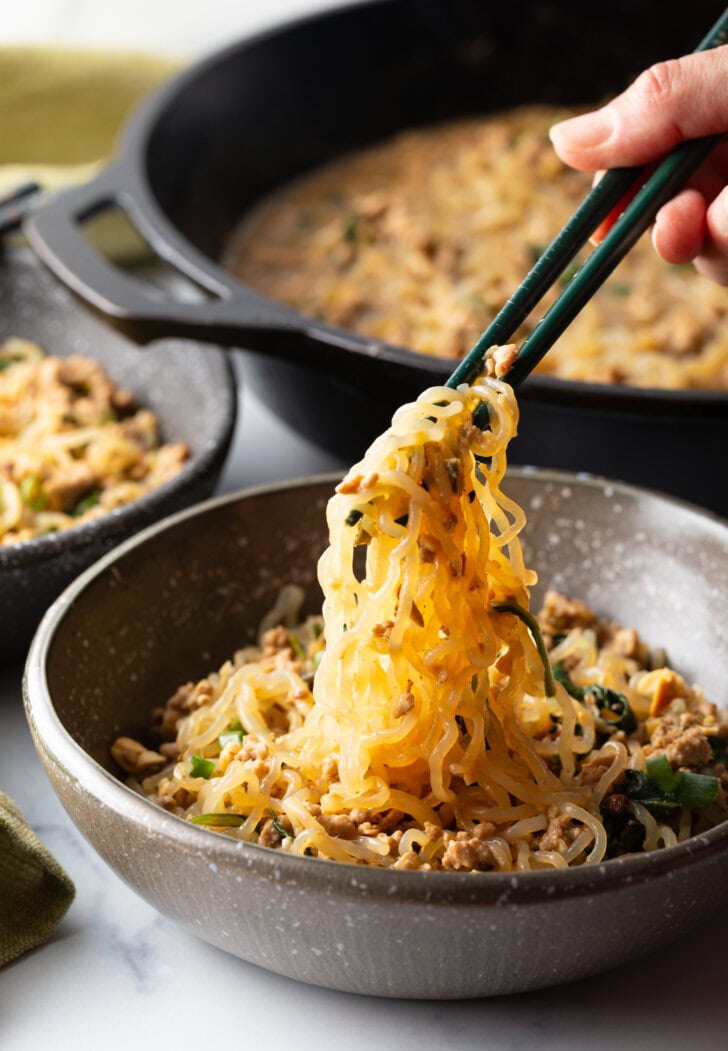 Spicy Szechuan noodles in a black bowl topped with green onion slices. Hand with set of chopsticks are grabbing a bite of noodle.