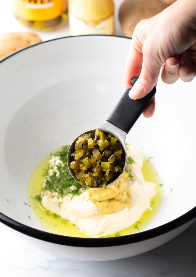 Hand holding measuring spoon with capers, adding to the mixing bowl.