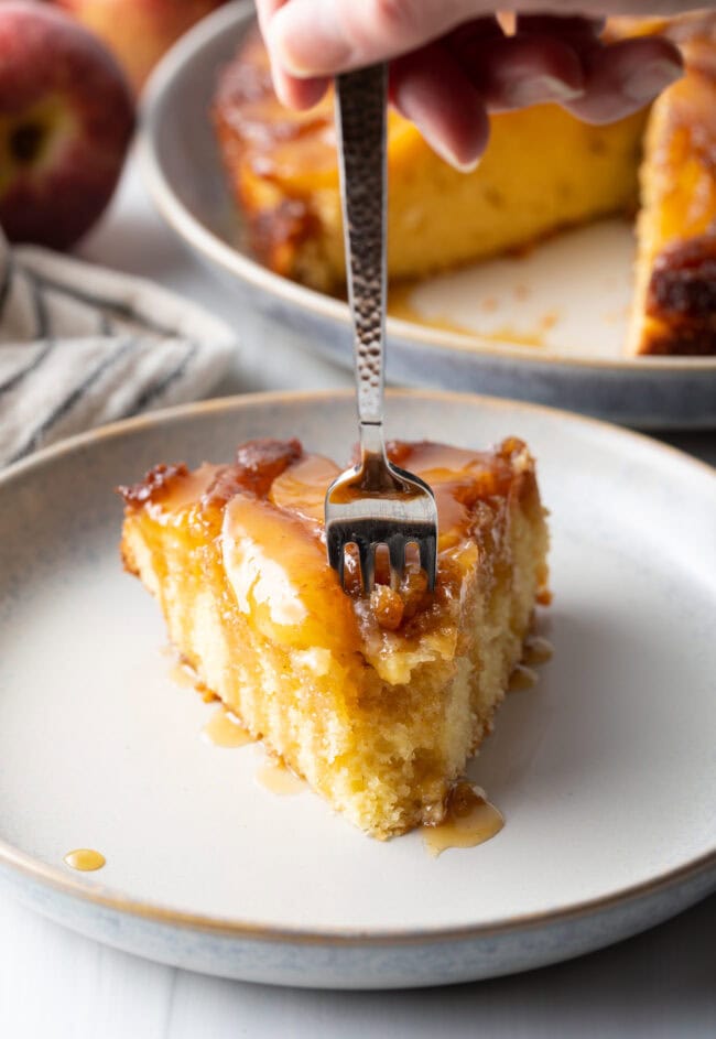 Fork taking a bite from slice of apple upside down cake on a white plate.