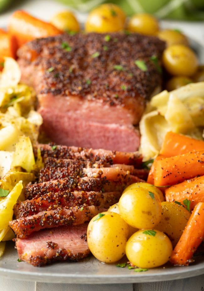 Sliced corned beef on a platter next to cabbage, potatoes and carrots. 