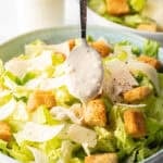 Spooning homemade Caesar dressing over a salad made with chopped romaine, croutons, and parmesan cheese.