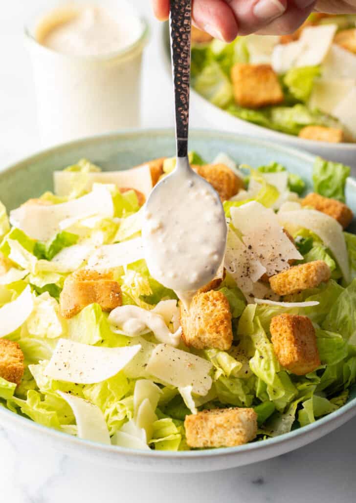 Spooning homemade Caesar dressing over a salad made with chopped romaine, croutons, and parmesan cheese.