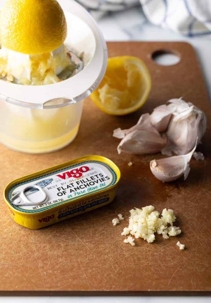 Ingredients on a cutting board: Container with fresh squeezed lemon, metal tin of anchovies, minced garlic and bowl garlic cloves.