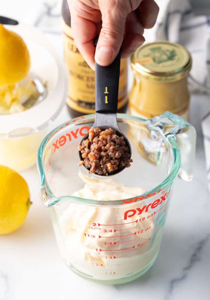 Adding a tablespoon of chopped anchovies to a glass measuring pitcher with mayonnaise.