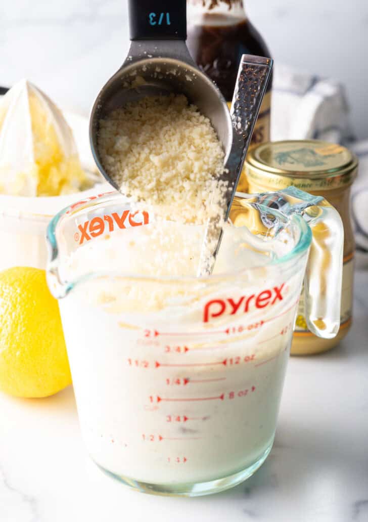 Adding shredded parmesan from a metal measuring cup into a glass pitcher with white dressing.