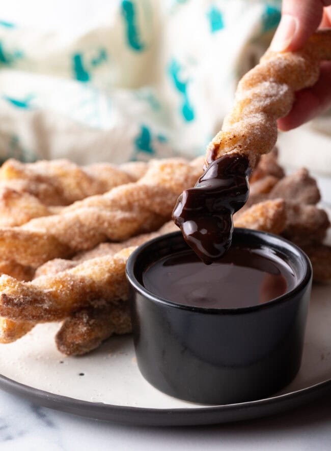 Hand dipping crunchy twist with sugar and cinnamon into a ramekin of chocolate sauce.