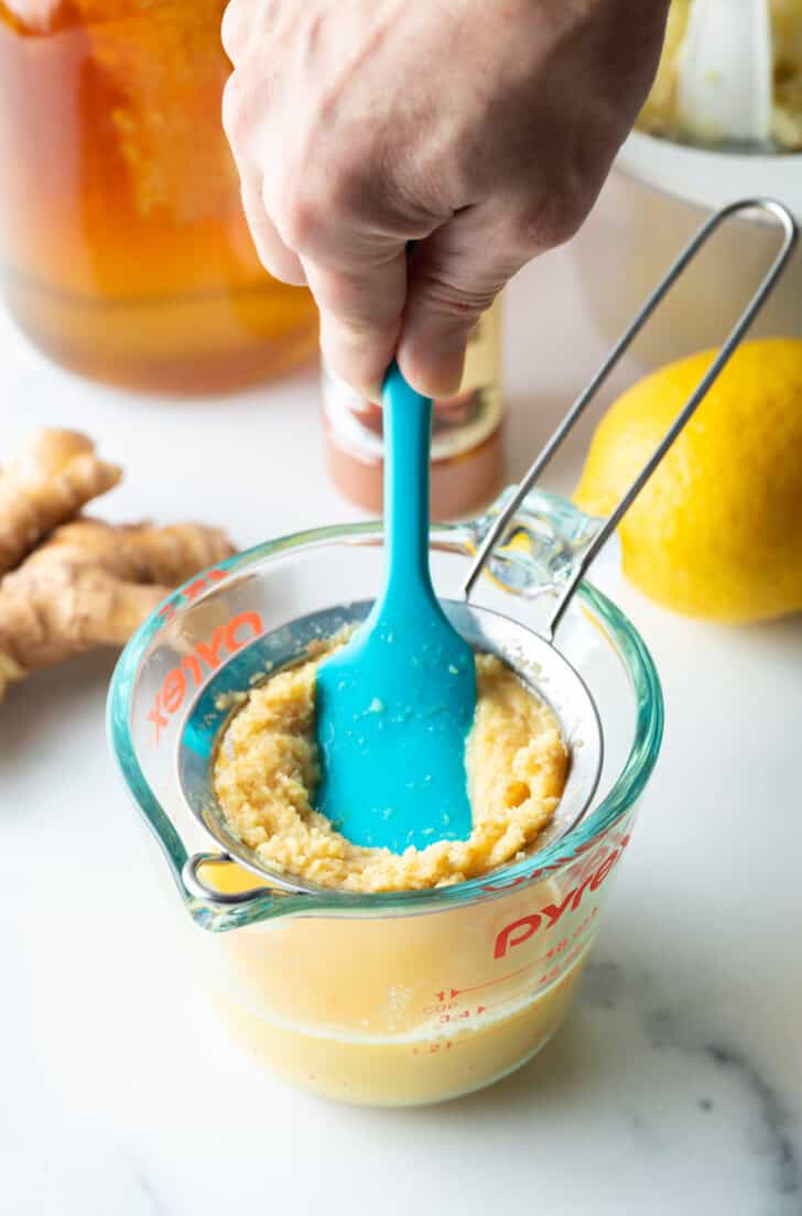 Using a blue spatula to push pureed ginger through a sieve.