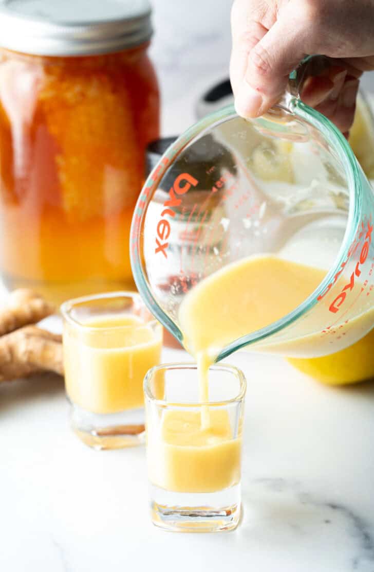 Pouring yellow liquid from a glass measuring pitcher into two shot glasses.