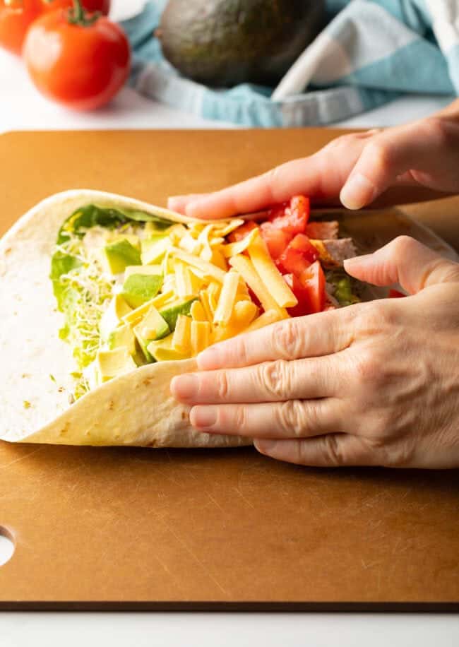 Two hands holding ingredients into the loaded tortilla while starting to roll into a wrap.