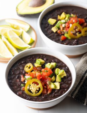 Two white bowls of black bean soup topped with slices of jalapenos, chopped avocado, and pico de gallo.