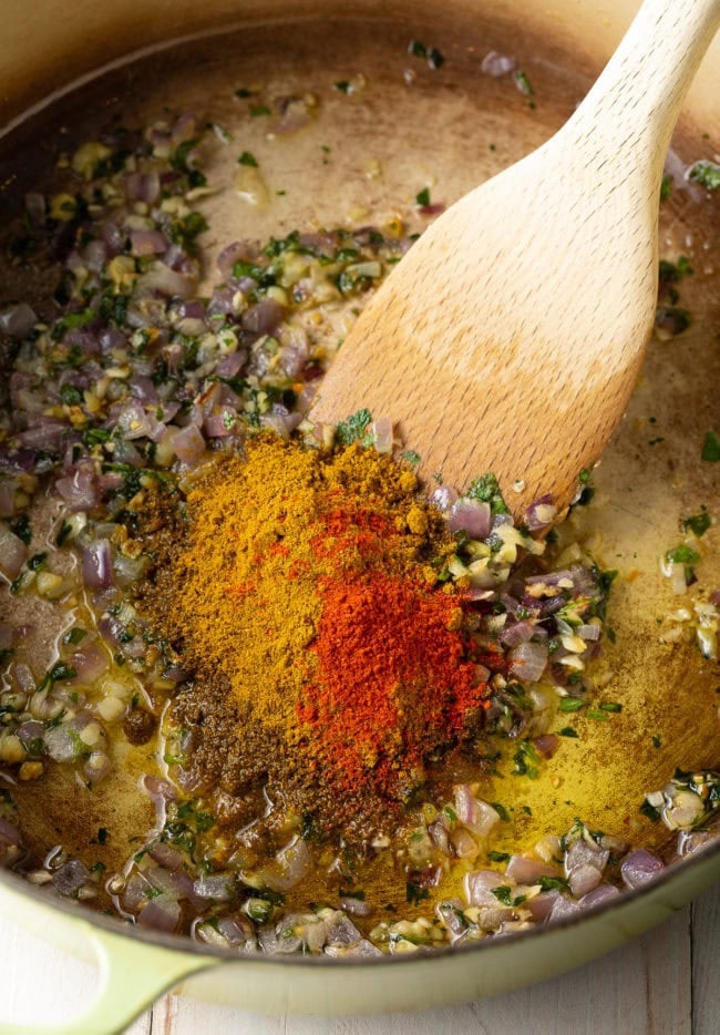 Spices and aromatics in a dutch oven being sautéed and stirred with a wooden spoon. 