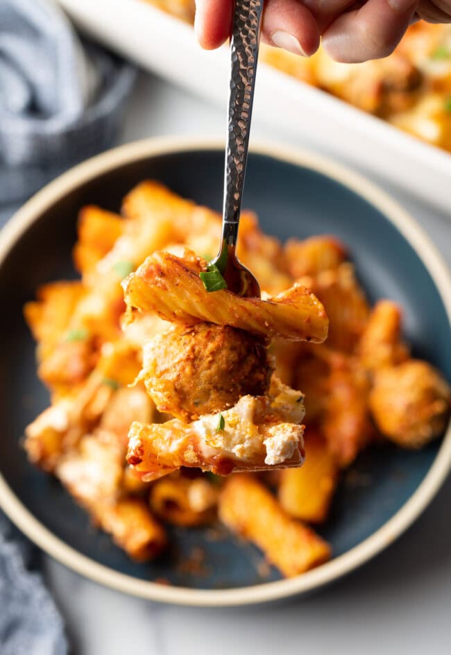 Dark gray plate with a generous portion of baked meatball casserole with pasta and cheese. Fork is holding a bite to camera.