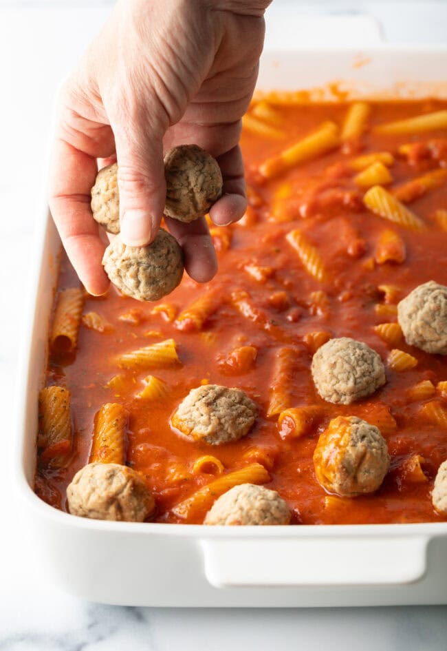 Hand adding frozen meatballs to baking dish with marinara and pasta.