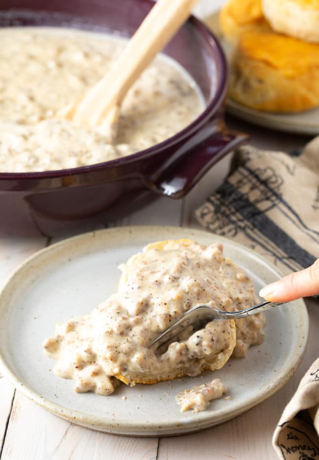 Hand cutting into a biscuit smothered with spicy sausage gravy on a plate. 