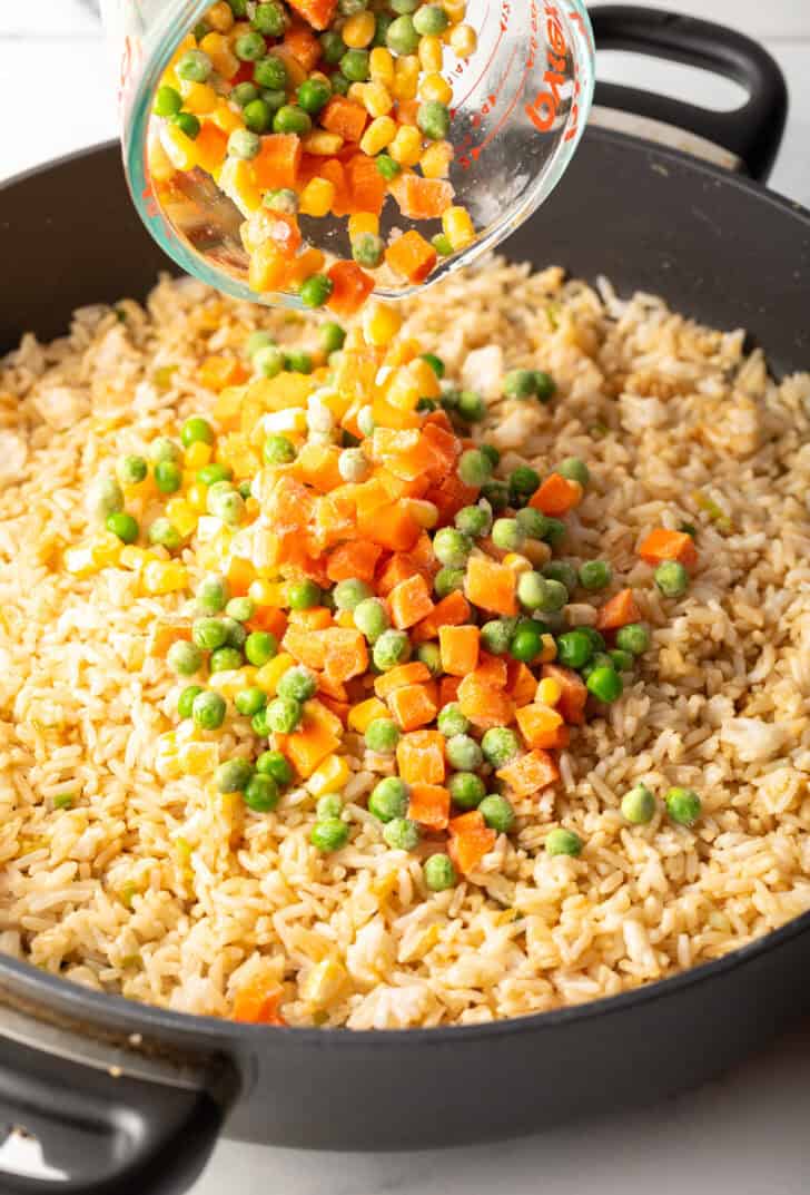 Adding frozen carrots, peas, and corn from a glass mixing cup to the rice in a skillet.