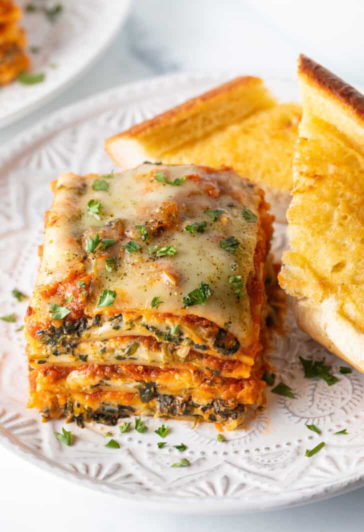 Square of cheesy spinach lasagna on a white plate with two slices of garlic bread.