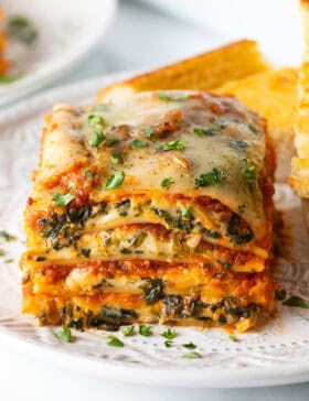 Square of cheesy spinach lasagna on a white plate with two slices of garlic bread.