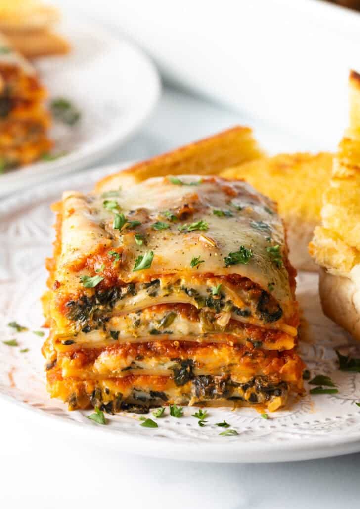 Square of cheesy spinach lasagna on a white plate with two slices of garlic bread.