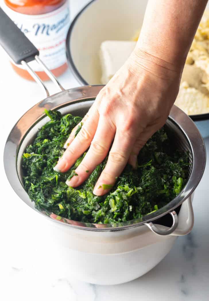 Hand pushing water out of spinach through a sieve.