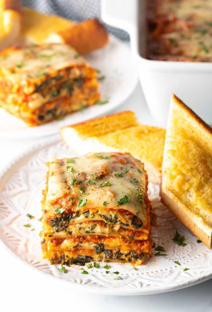 Square of cheesy spinach lasagna 
 recipe on a white plate with two slices of garlic bread.