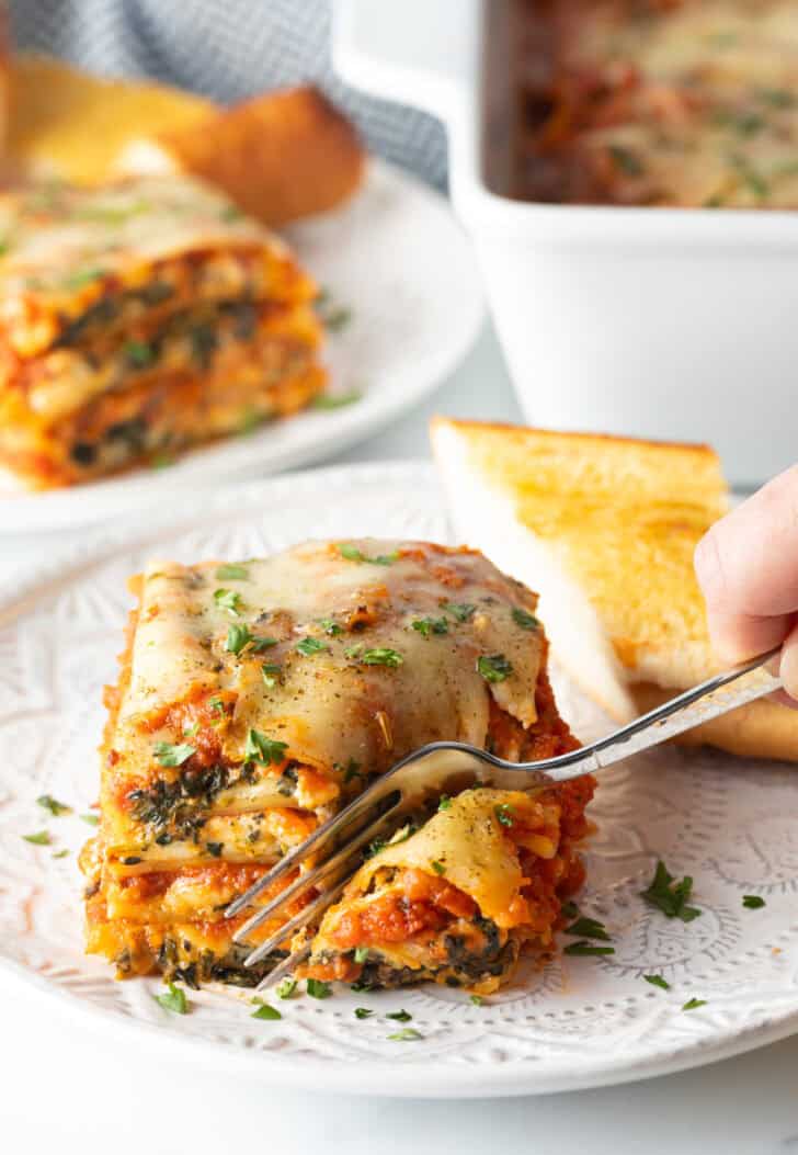 Hand with a fork, taking a bite from a serving of lasagna on a white plate.