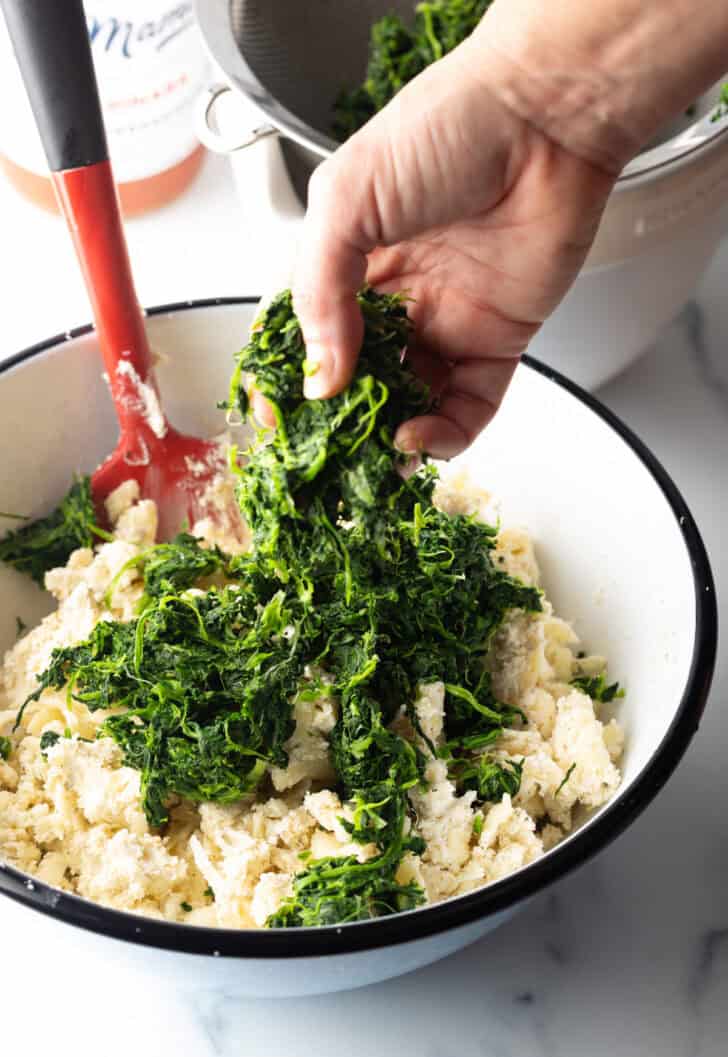 Hand adding frozen and thawed spinach to a mixing bowl with cheese.