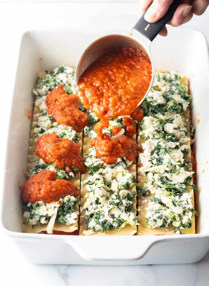 Pouring tomato sauce from a measuring cup, over layers of lasagna noodles topped with shredded cheese and spinach.