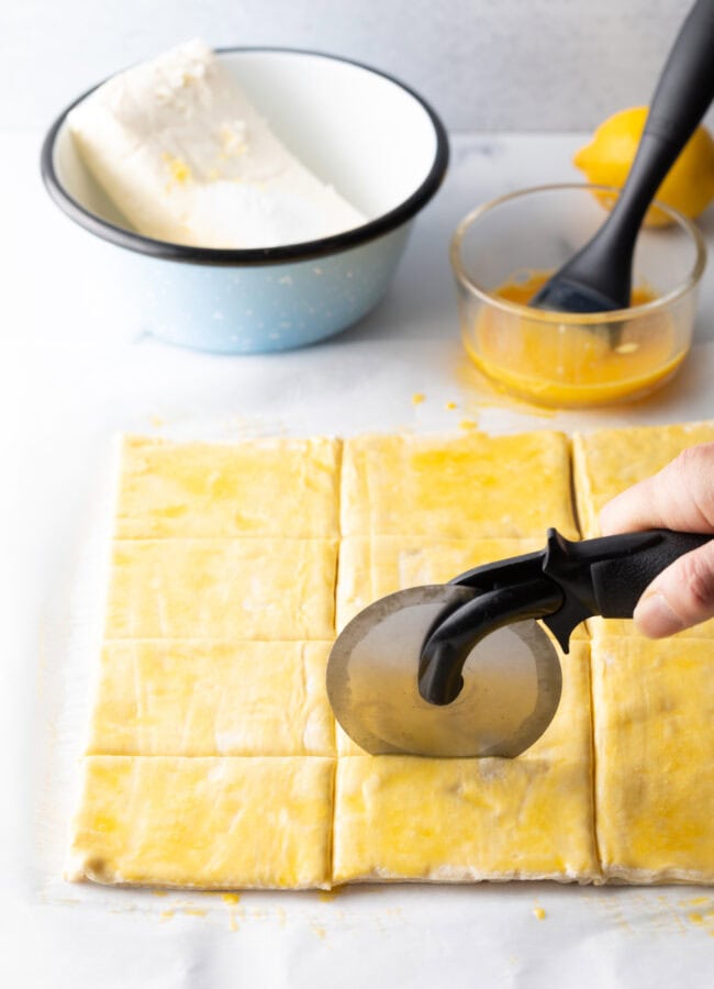 Hand using a pizza cutter to slice a sheet of puff pastry into rectangles.