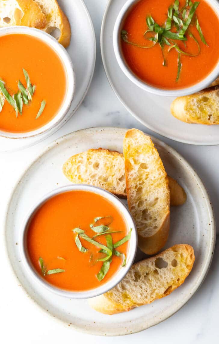 Top down view a bowl of tomato soup on a white plate, surrounded by three slices of baguette.