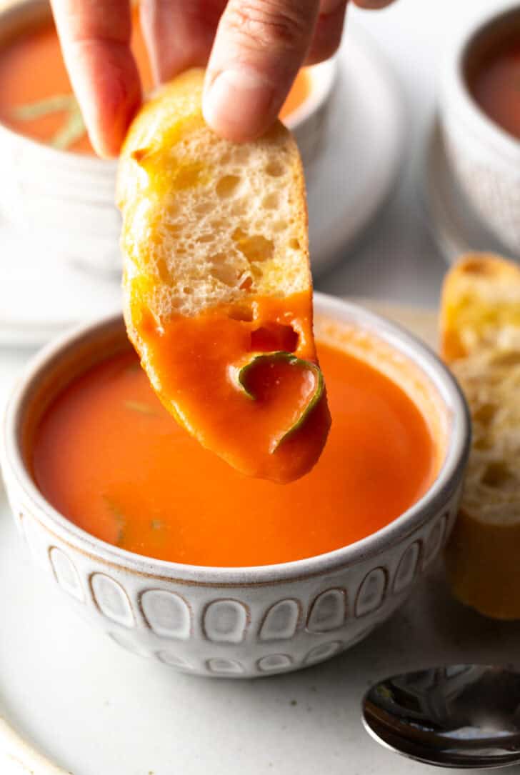 Hand dipping a slice of baguette into rich tomato soup.