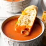 White bowl of tomato soup with a slice of baguette in the soup.