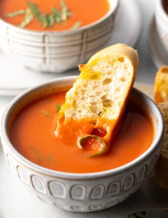 White bowl of tomato soup with a slice of baguette in the soup.