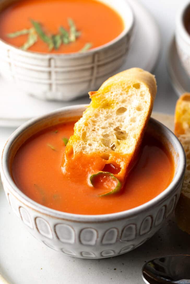 White bowl of tomato soup with a slice of baguette in the soup.