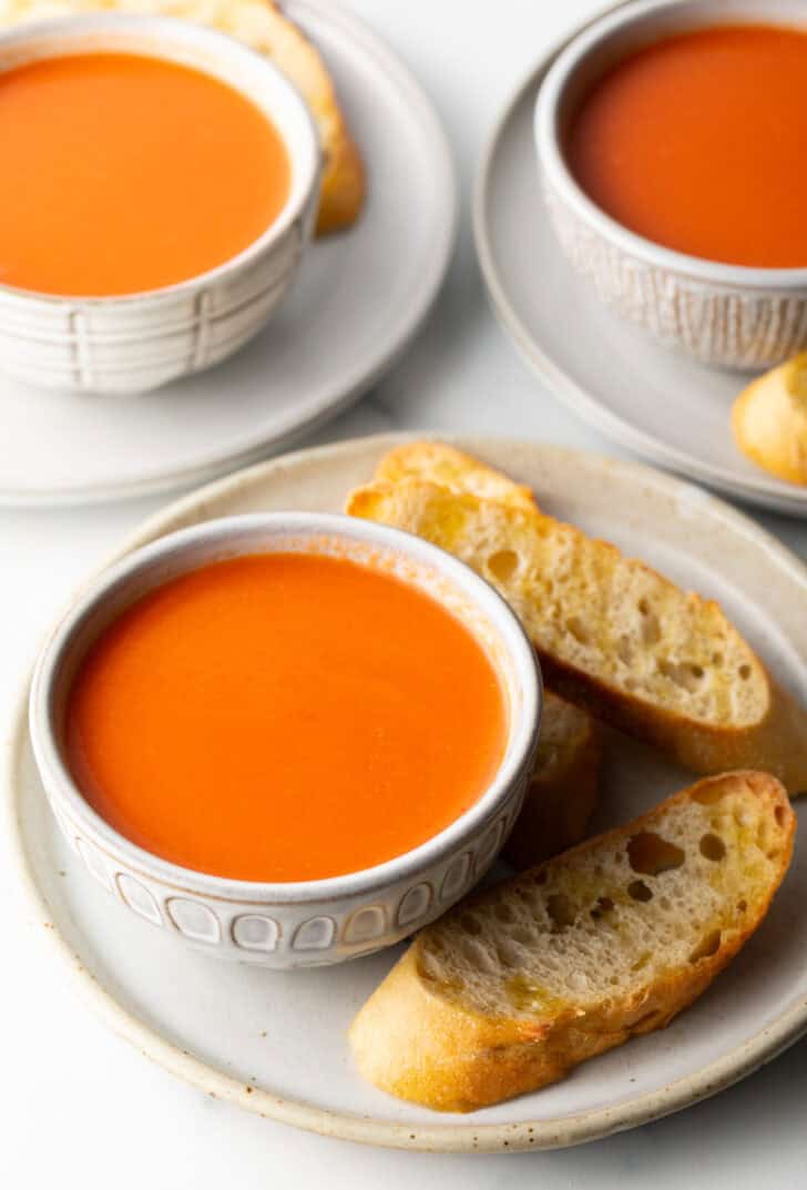 Bowl of tomato soup on a white plate, surrounded by three slices of baguette.