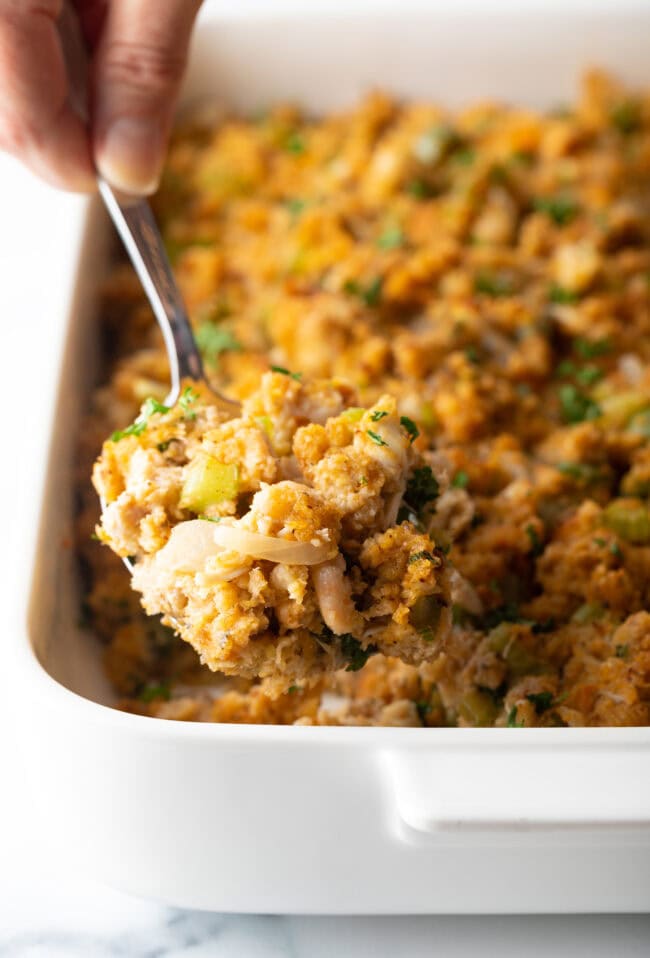 Metal spoon holding a serving of stuffing to camera. In the background is the white baking dish with seafood dressing.